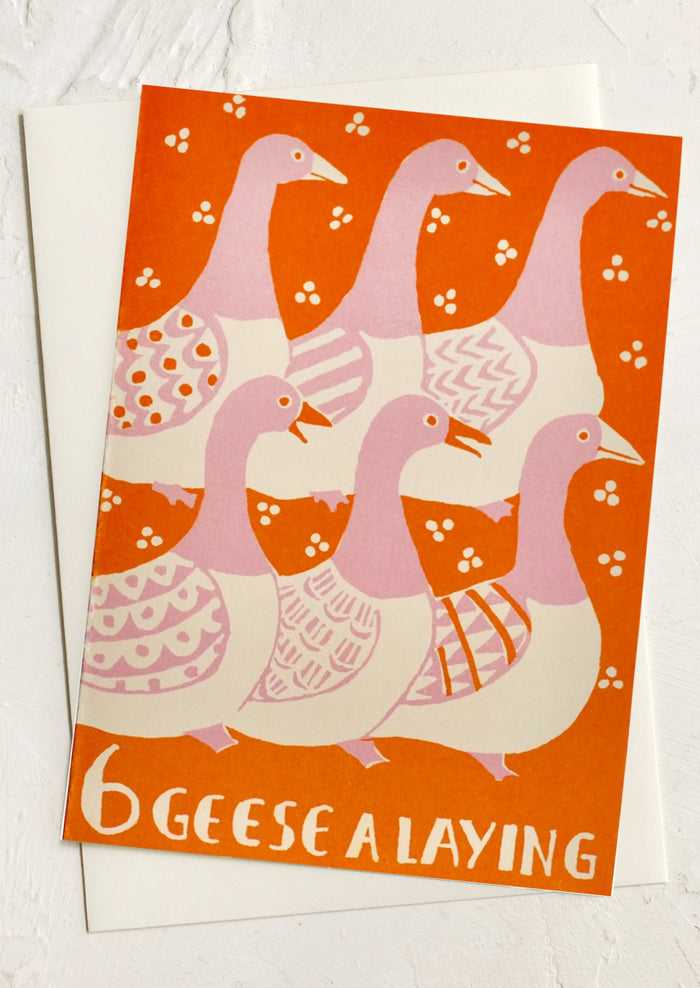 A vintage-style greeting card featuring an illustration of six pink and cream geese on an orange background, with the text '6 GEES E A LAYING' at the bottom. The card is displayed on top of a white envelope against a light textured background.
