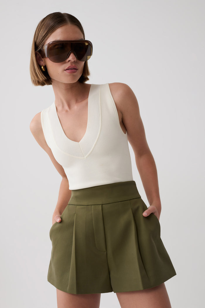 A model wearing a cream sleeveless V-neck knit top and olive green pleated shorts, styled with oversized dark sunglasses, against a light gray studio background.