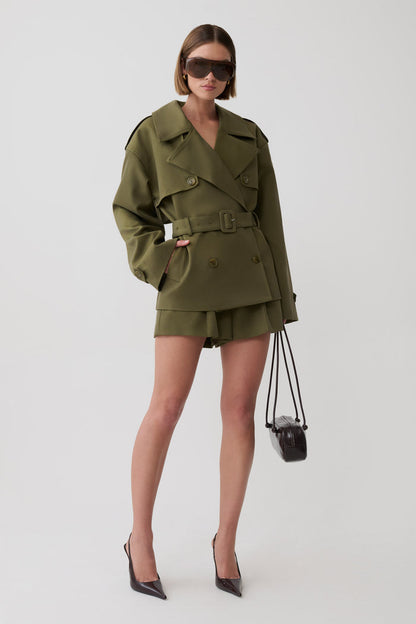 A model wearing an oversized olive green military-style trench jacket and matching shorts with a belt, styled with black pointed-toe heels and a small black shoulder bag against a light gray studio background.