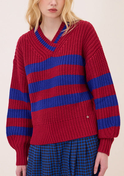 A model wearing a chunky knit V-neck sweater with horizontal red and blue stripes, paired with a blue and black checkered skirt, against a plain light beige background.