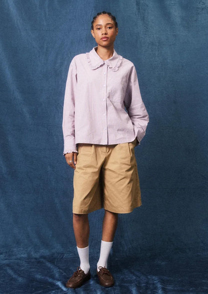 A model stands against a textured blue backdrop wearing a light pink long-sleeve button-up shirt with a ruffled collar, tan shorts, white socks, and brown loafers.