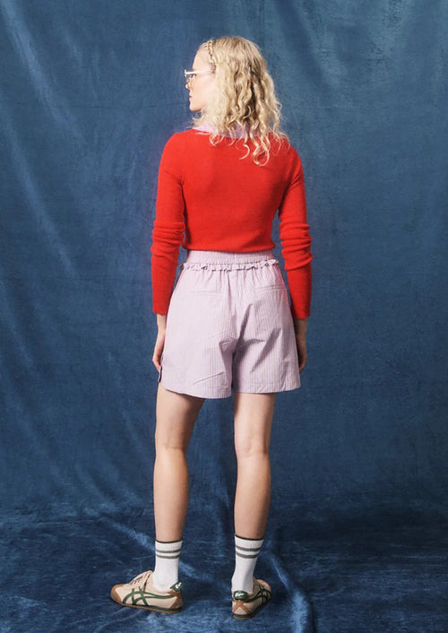 A woman stands with her back to the camera wearing a bright red long-sleeve top, light pink pleated shorts, striped socks, and sandals, posed against a textured blue fabric backdrop.