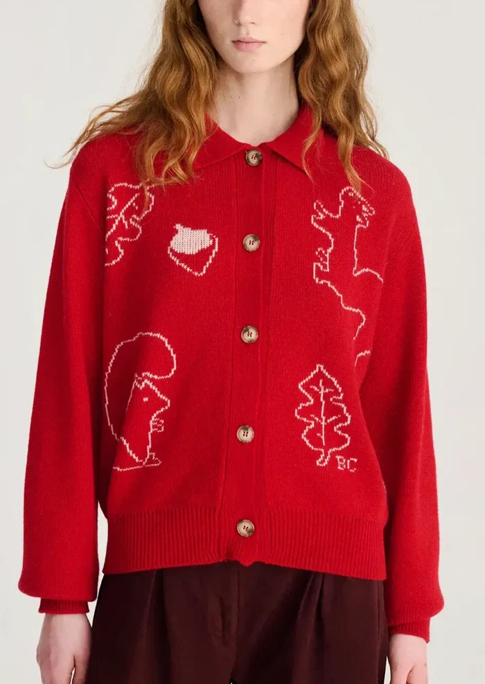 A red knit cardigan sweater with white knitted animal and leaf motifs, featuring gold buttons and a collar, worn by a model.