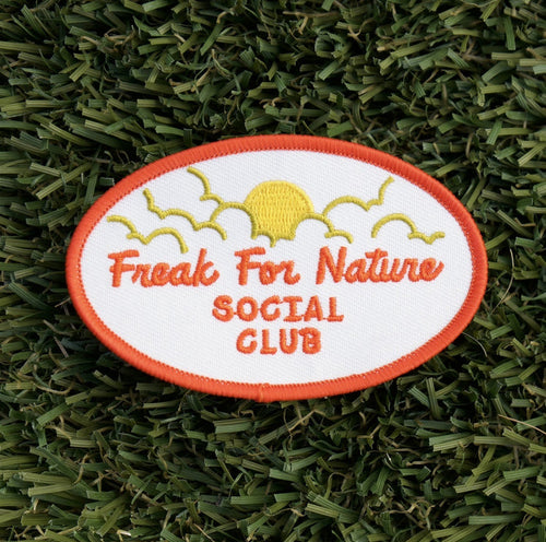 Oval-shaped iron-on embroidered patch with a white background and red words that say "Freak for nature social club" and a yellow sun and clouds.