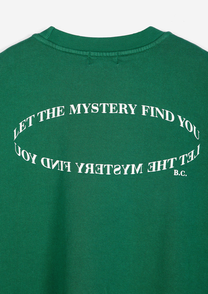 A close-up of a green crewneck sweatshirt showing the back, which features a white graphic text design that reads 'LET THE MYSTERY FIND YOU' in an oval shape, with 'B.C.